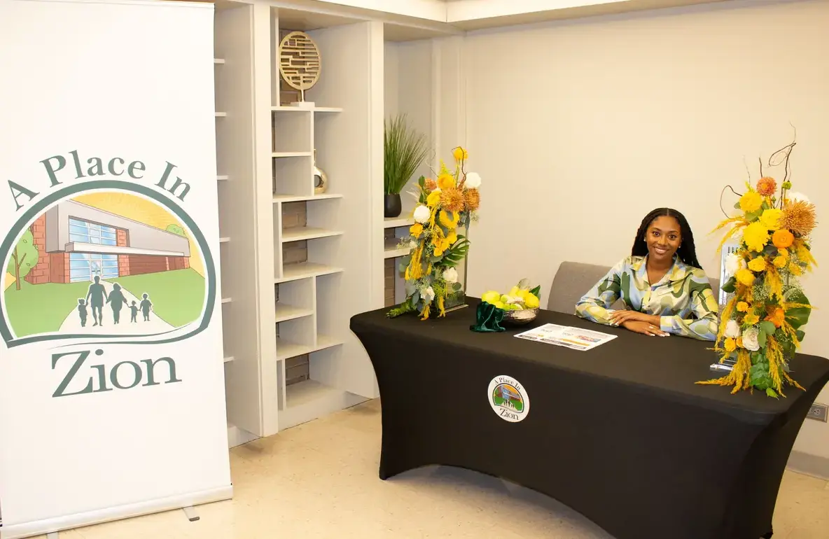 A beautiful young lady sitting at a sign-up table decorated with flowers beisde a standing banner that has the logo for A Place in Zion